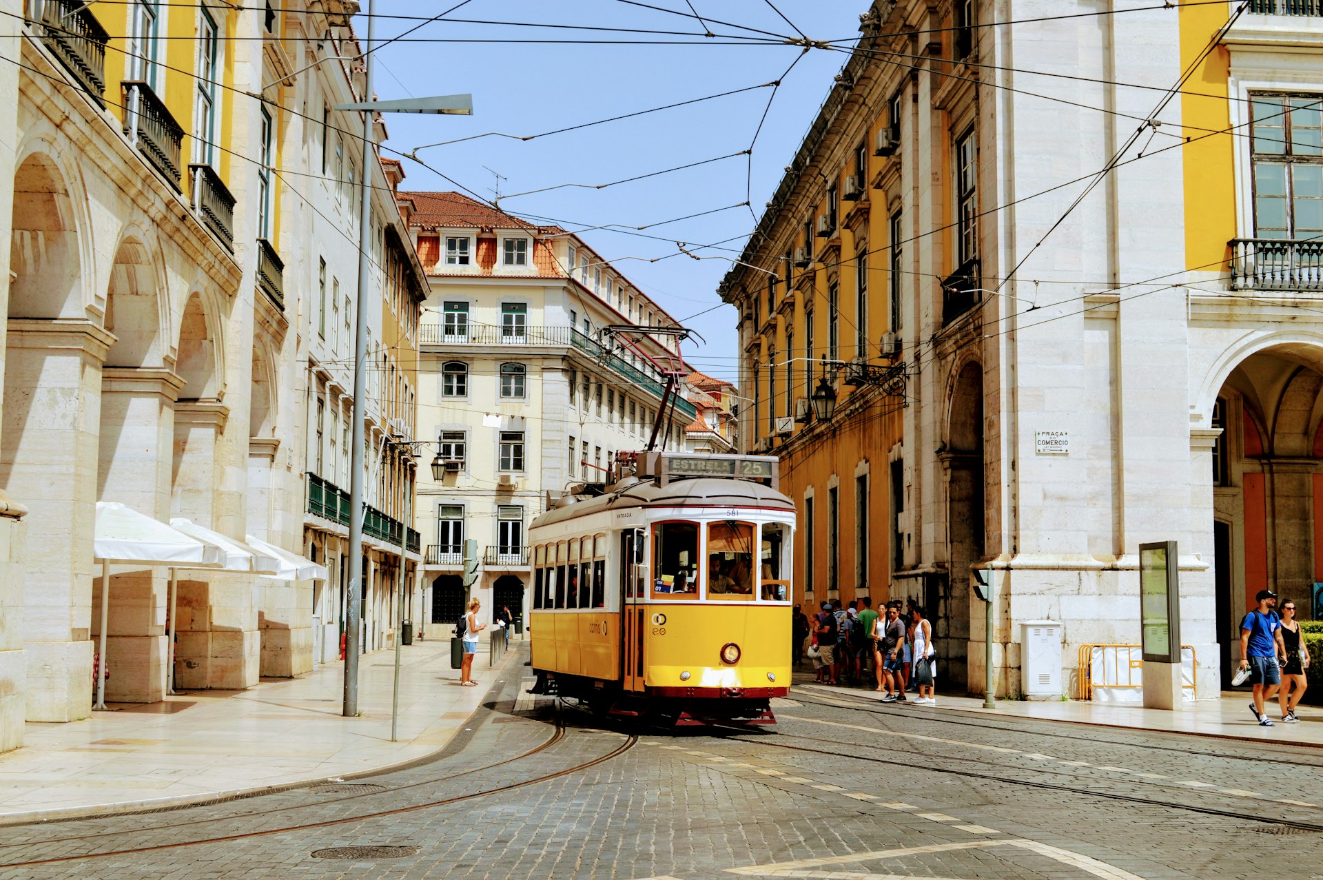 A yellow number 25 tram rounding the corner onto a cobbled street by Praca do Comercio in Lisbon